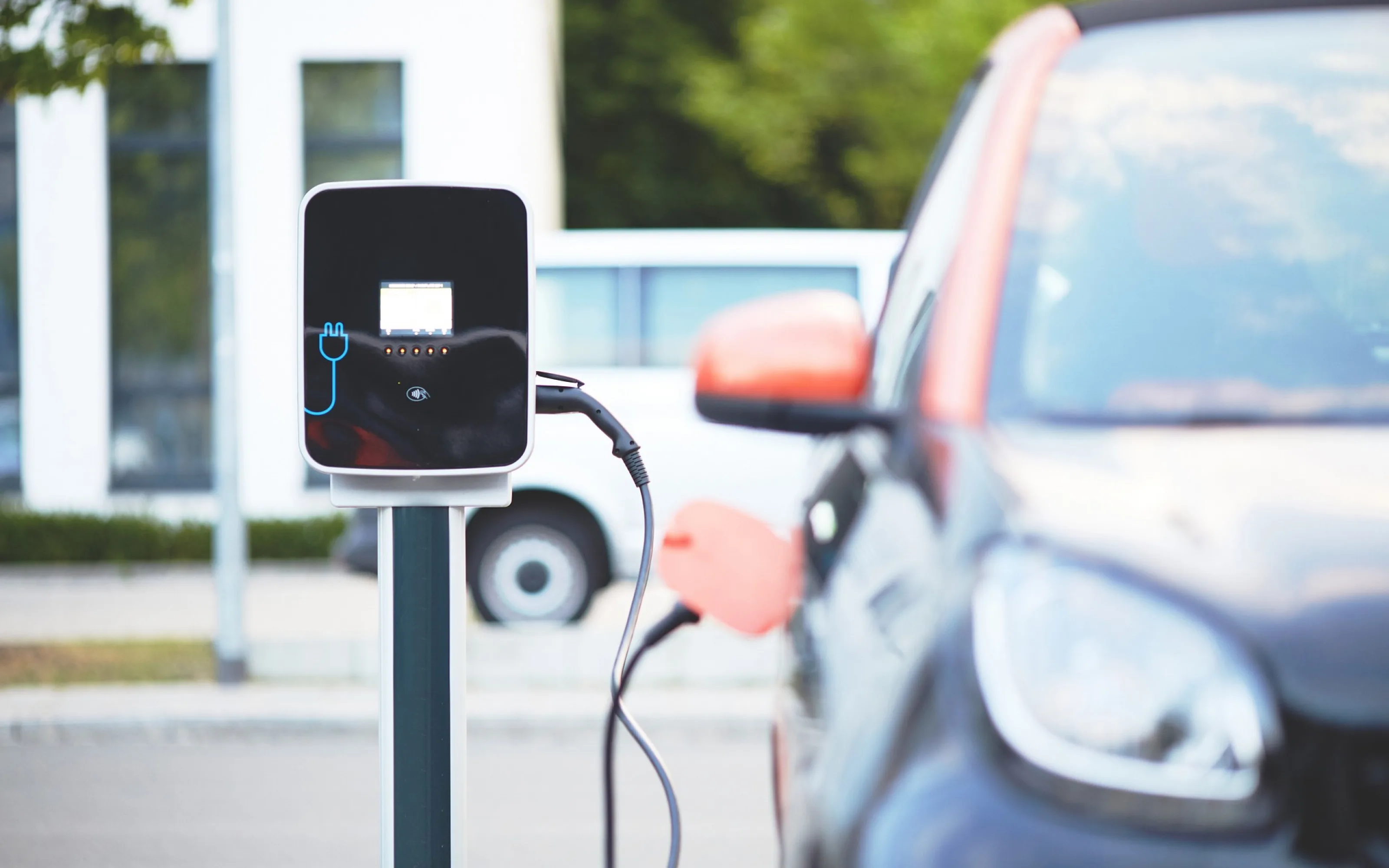 A charging station with a small screen and a blue plug icon, its cable plugged into a dark electric car with an orange mirror, blurred office windows, trees and a white van behind