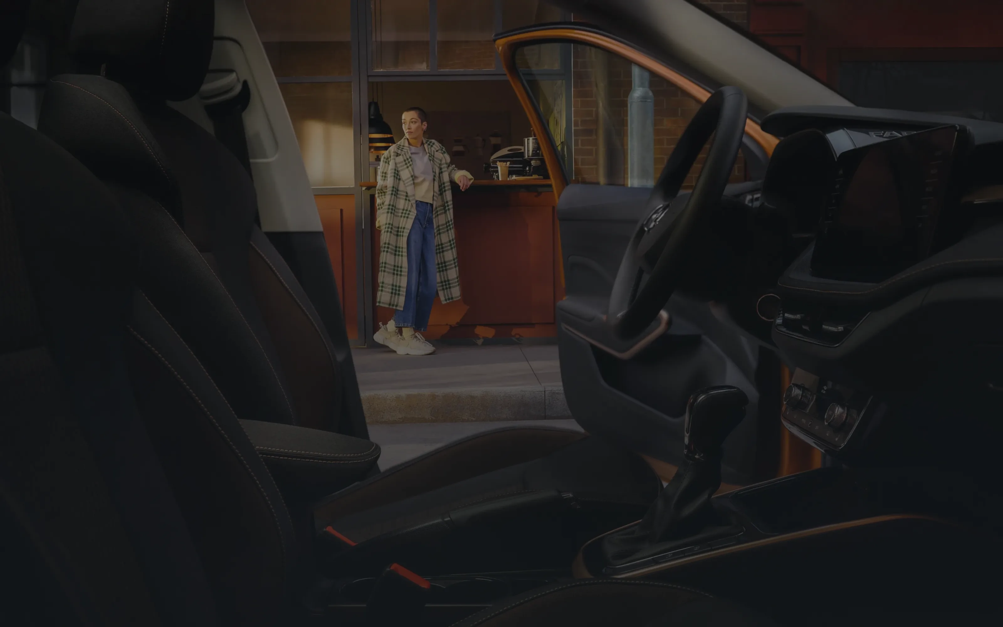 Seen from inside a car at night, past the seats and steering wheel: the open passenger door frames a woman in a green-checked coat and white sneakers leaning on a lit café counter, waiting for the car handover