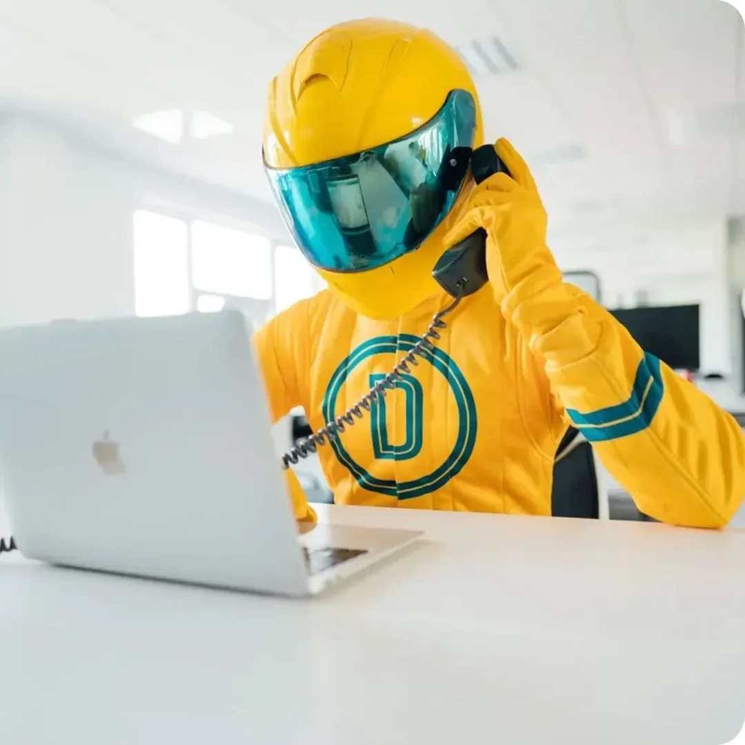 Maskot Driveramy ve žluté kombinéze a zrcadlové přilbě telefonuje u stolu s otevřeným notebookem