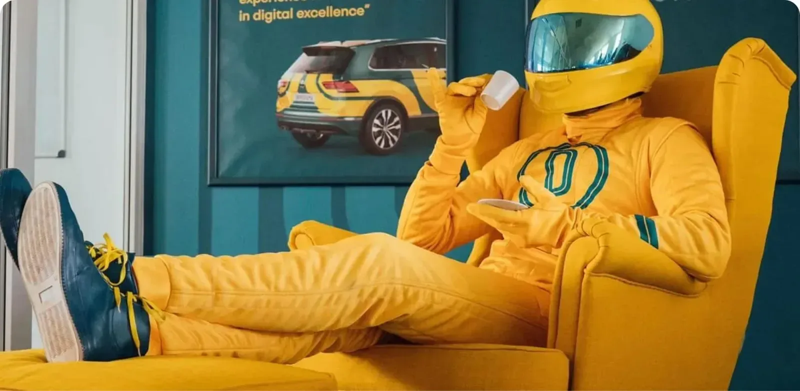 Maskot Driveramy ve žluté závodní kombinéze a zrcadlové přilbě se rozvaluje ve žlutém křesle pod zarámovaným plakátem se žlutým autem