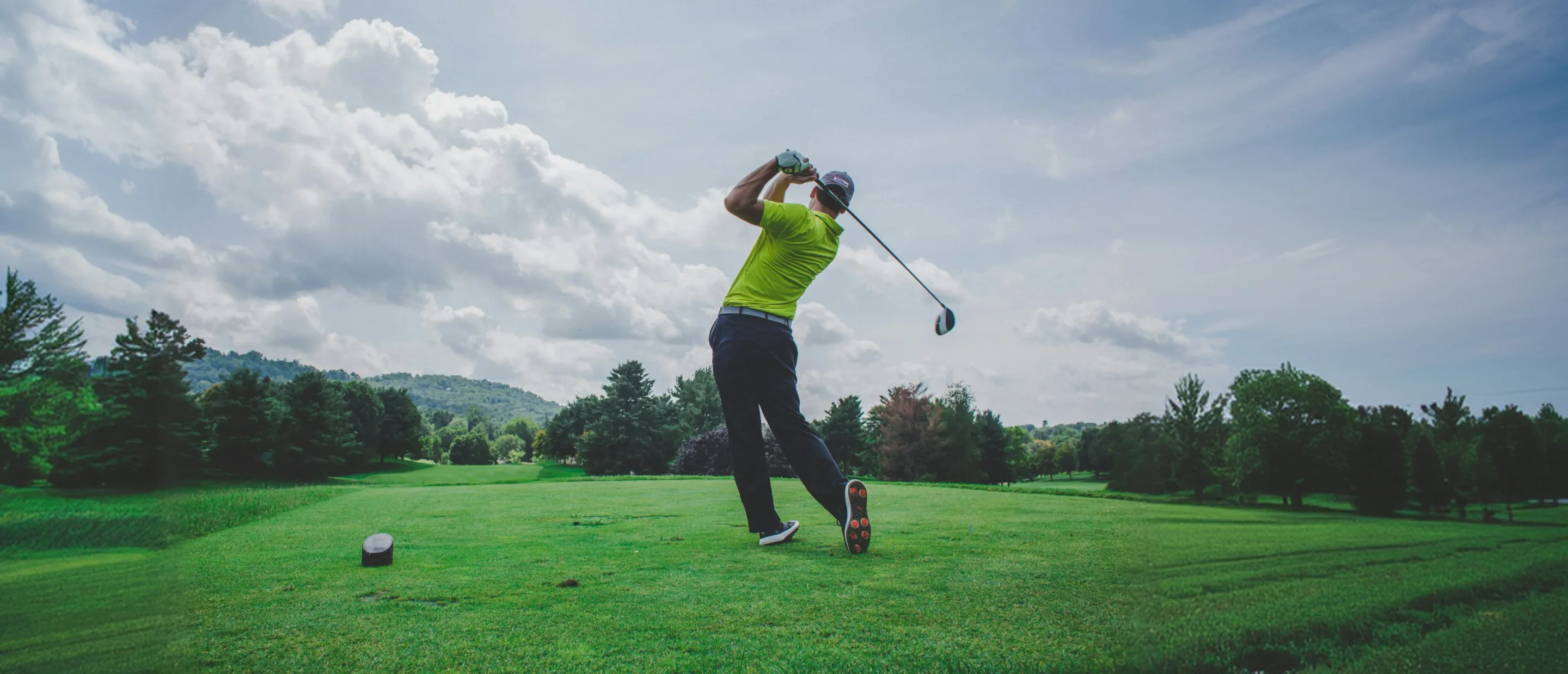 A golfer in a lime polo at the end of his drive on a tee box, the club swung over his shoulder, trees and a cloudy sky behind him