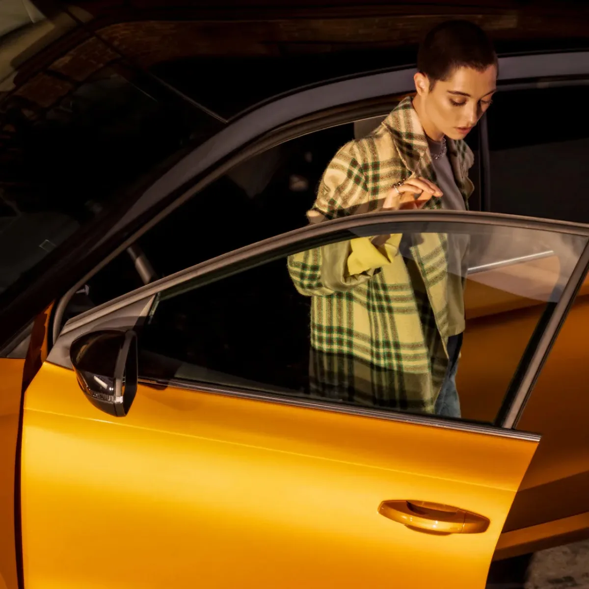 A woman in a checked shirt looking down as she opens the door of a yellow car in a dim garage