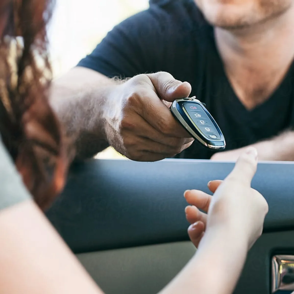 A man in a black T-shirt handing a car key fob through the car window to a woman's open hand