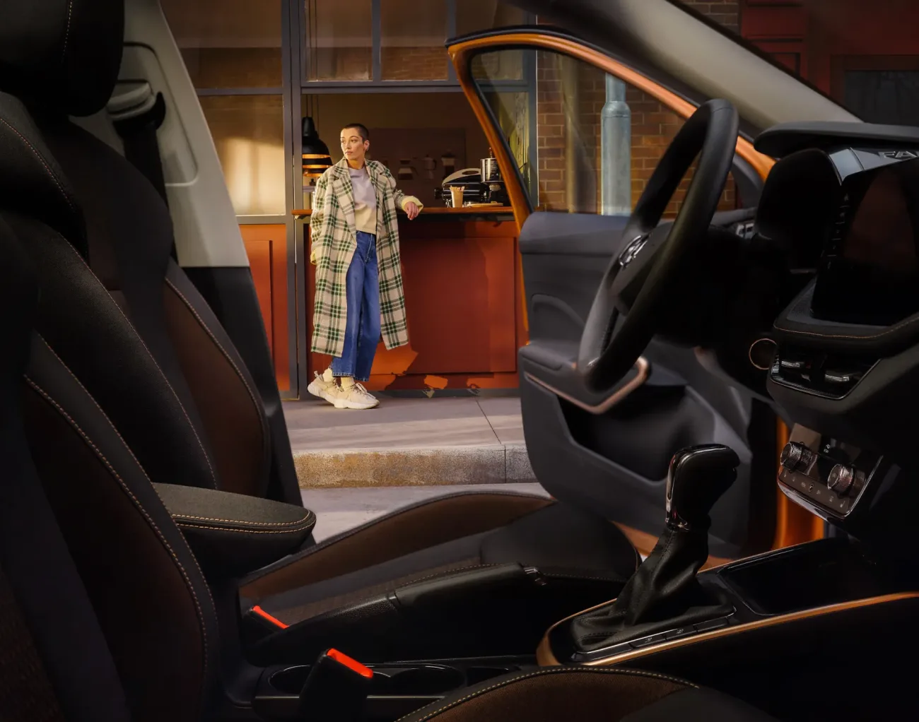 Seen from inside a car past the seats and gear stick: through the open passenger door a woman in a green-checked coat leans on a lit café counter, waiting for the handover