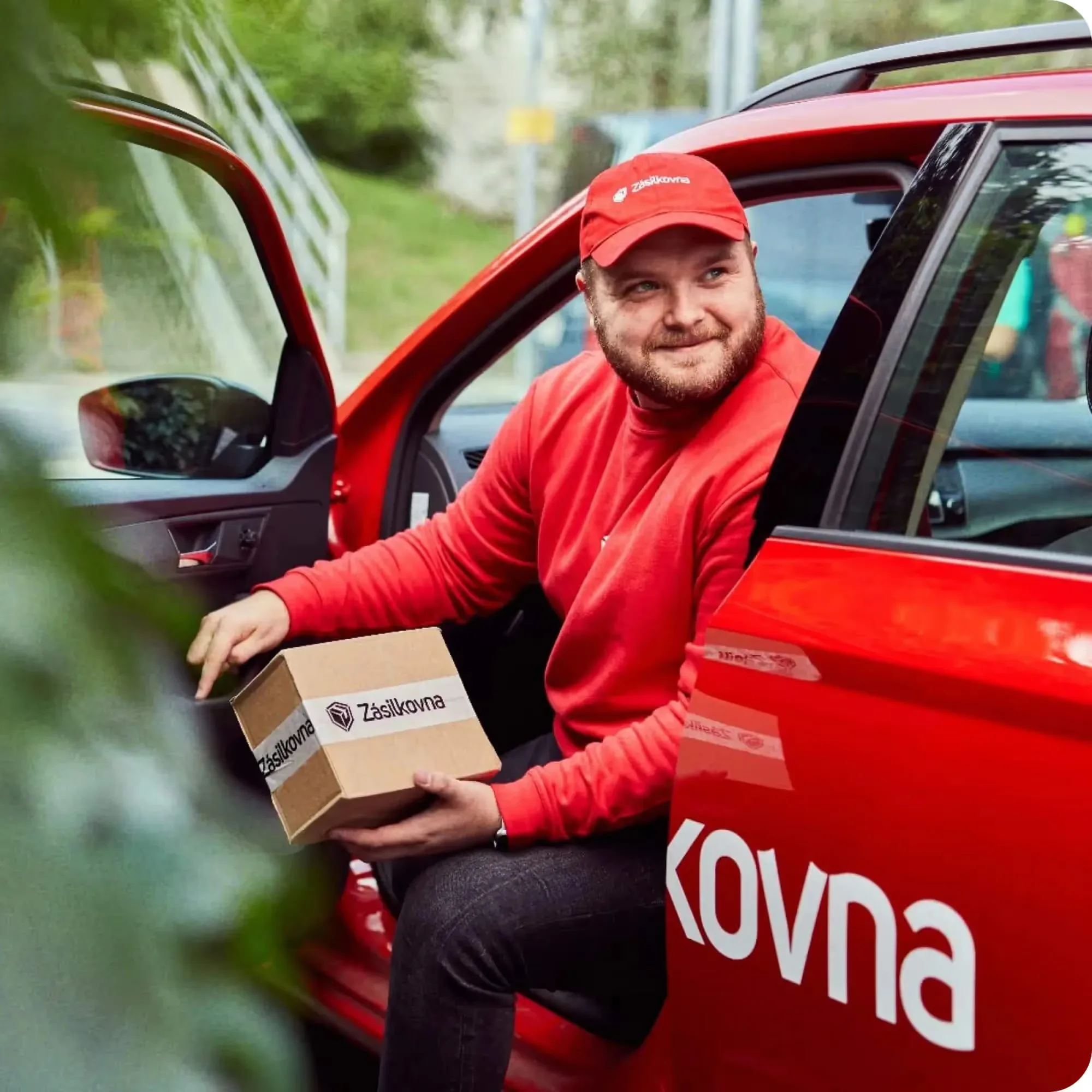A smiling Zásilkovna courier in a red cap and sweatshirt stepping out of a red delivery car with a cardboard parcel sealed with Zásilkovna tape