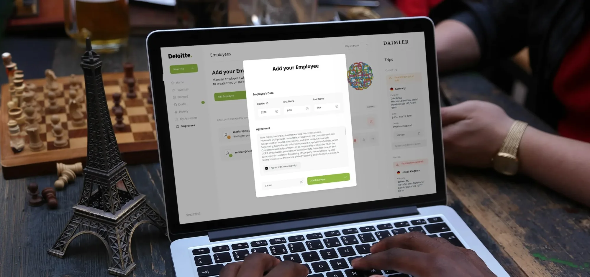 Ruce píšící na notebooku u dřevěného stolu v kavárně vedle šachovnice, piva a figurky Eiffelovy věže; na obrazovce je stránka Employees v compliance aplikaci s dialogem Add your Employee – ID zaměstnance, jméno a příjmení, souhlas se zpracováním osobních údajů a tlačítko Add Employee – a panel Trips s cestou Daimler do Německa