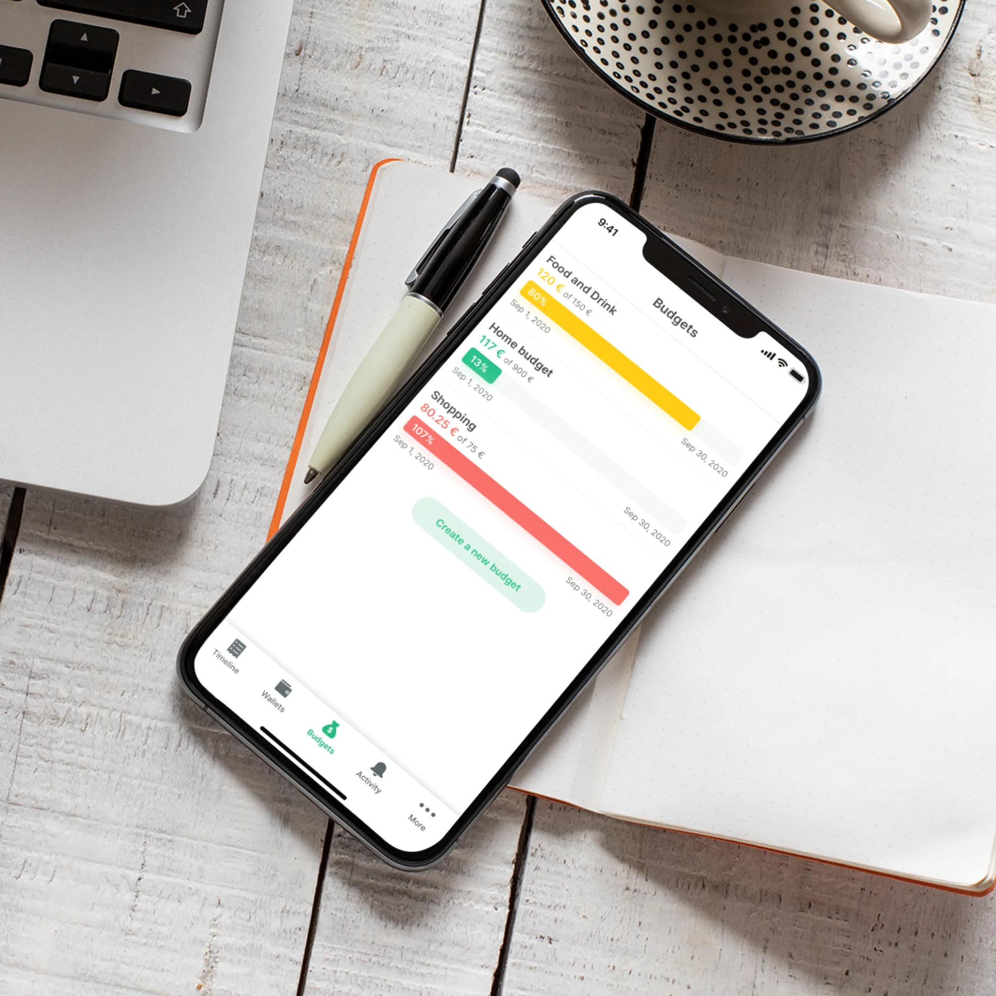 An iPhone lying on a white wooden desk beside a laptop, an open orange notebook, a pen and a dotted cup, showing the Spendee Budgets screen — Food and Drink 80%, Home budget 13% and Shopping 107% progress bars